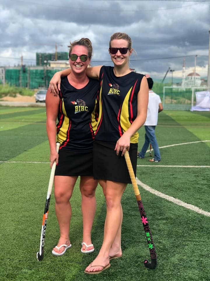 Two women are standing next to each other on a field holding hockey sticks.