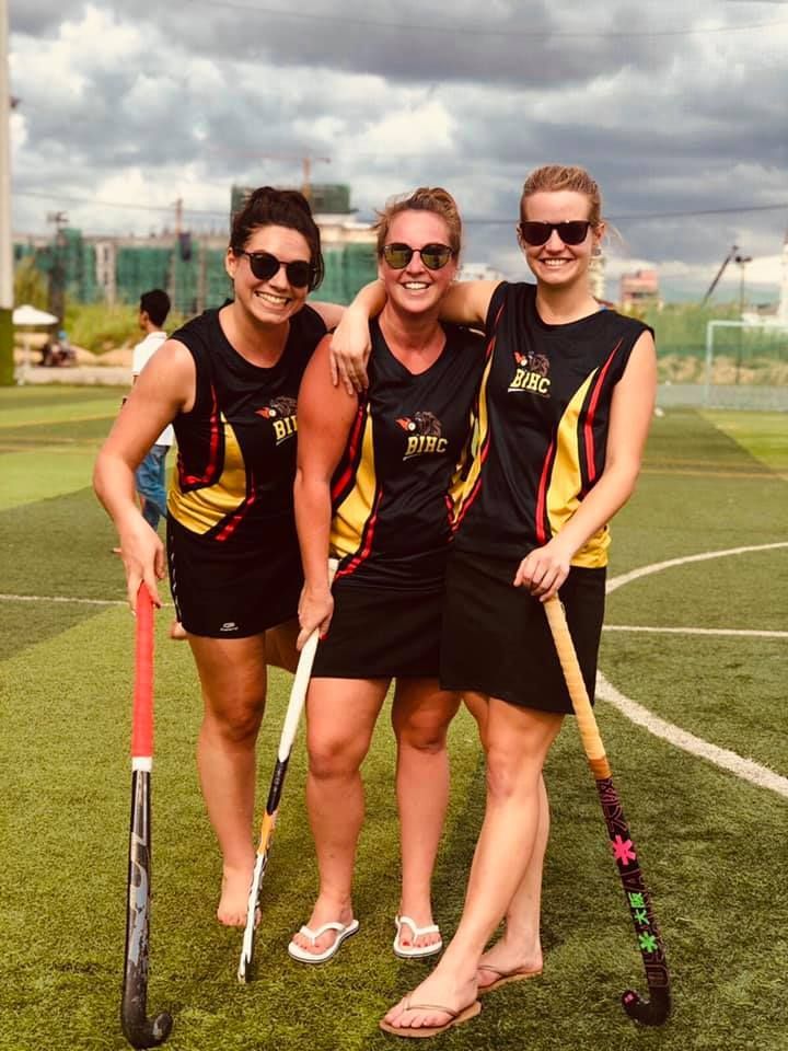 Three women are standing on a field holding hockey sticks.