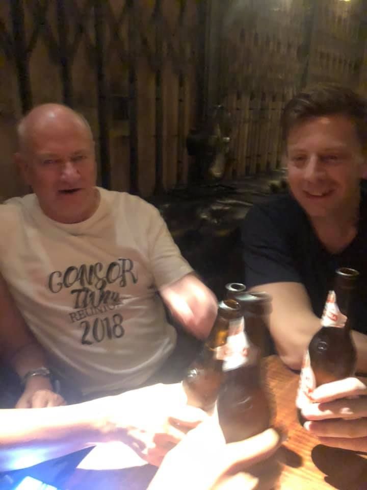 Two men are sitting at a table with bottles of beer and one of them is wearing a shirt that says cancer time 2018