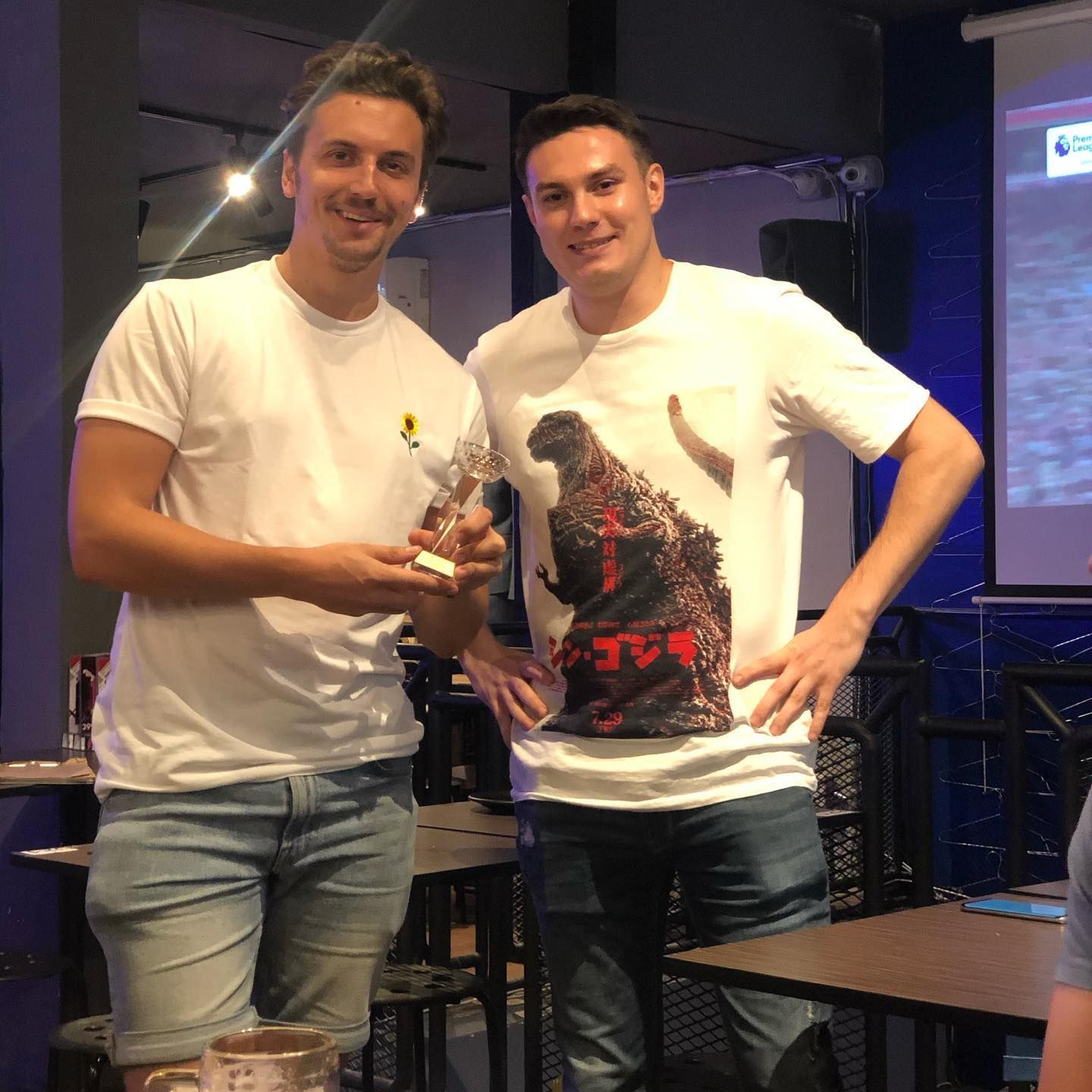 Two men are standing next to each other in a room and one has a godzilla shirt on