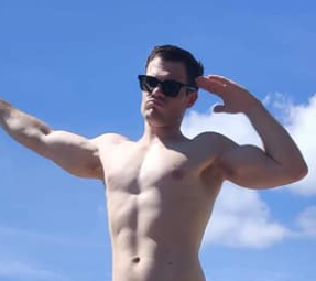 A shirtless man wearing sunglasses is standing on a beach with his arms outstretched.