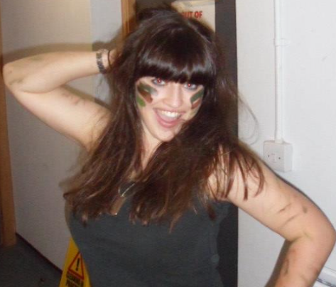 A woman with camouflage paint on her face stands next to a yellow wet floor sign