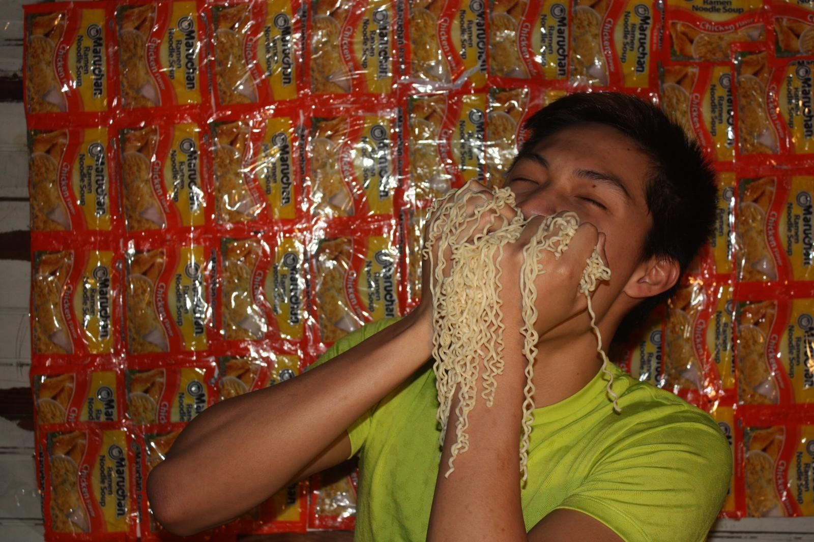A young man is eating noodles in front of a wall of noodles