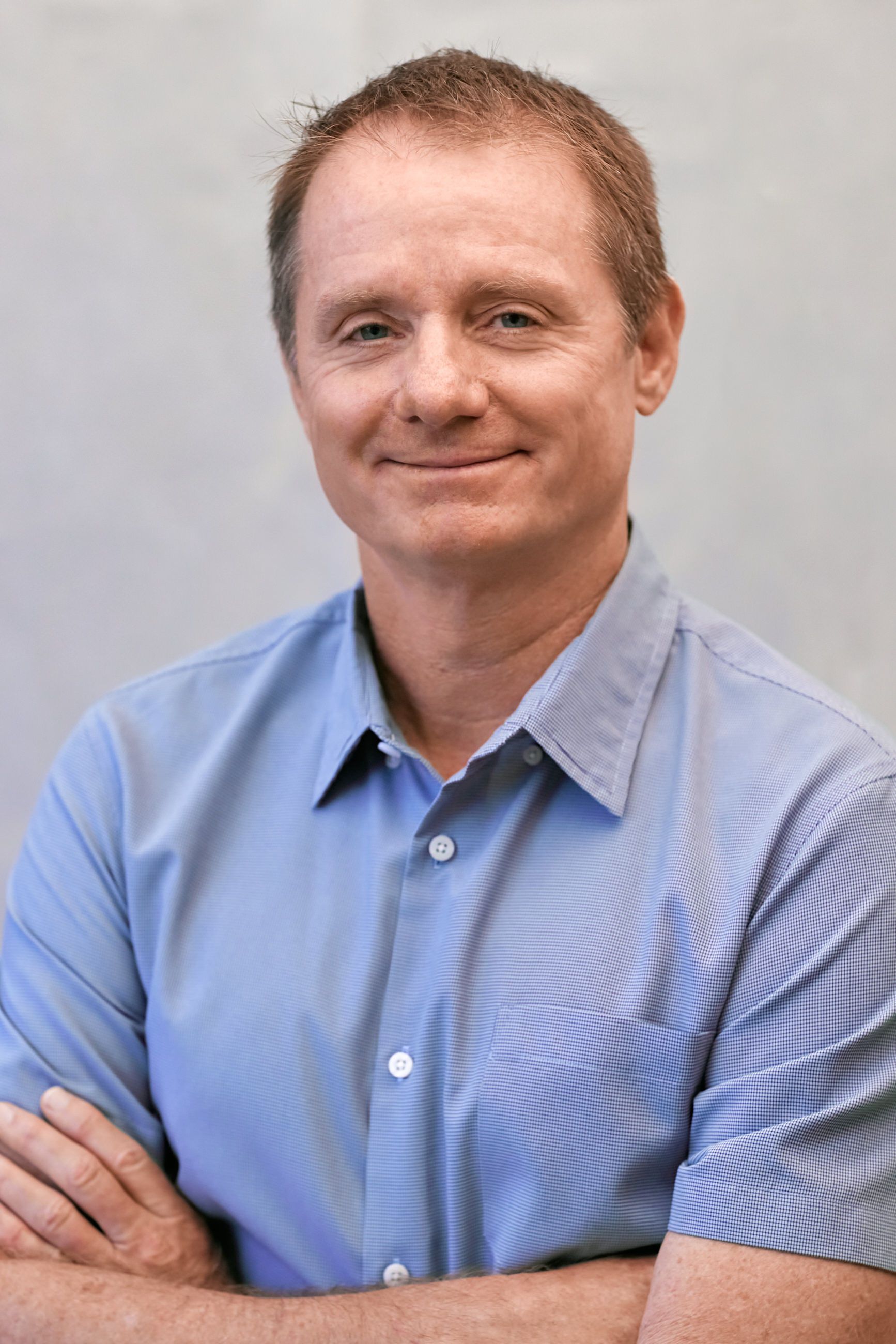 A man in a blue shirt is smiling with his arms crossed.
