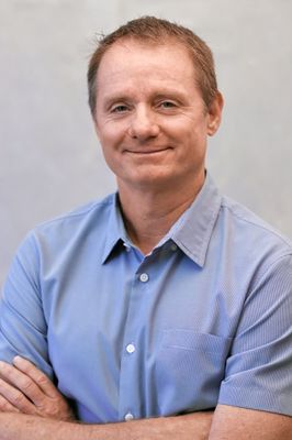 A man in a blue shirt is smiling with his arms crossed.