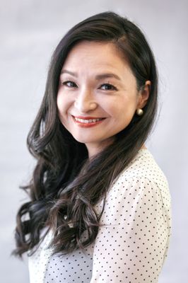 A woman with long hair is smiling and wearing a white polka dot shirt.