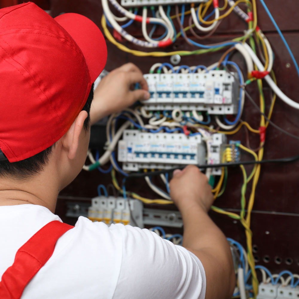 Electrician in a red cap working on an electrical panel, wiring visible.
