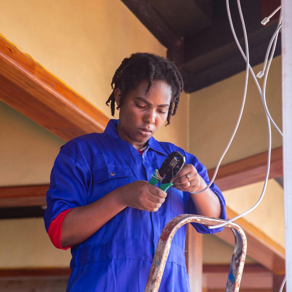 Woman in blue jumpsuit crimping wires on a ladder near ceiling beams.