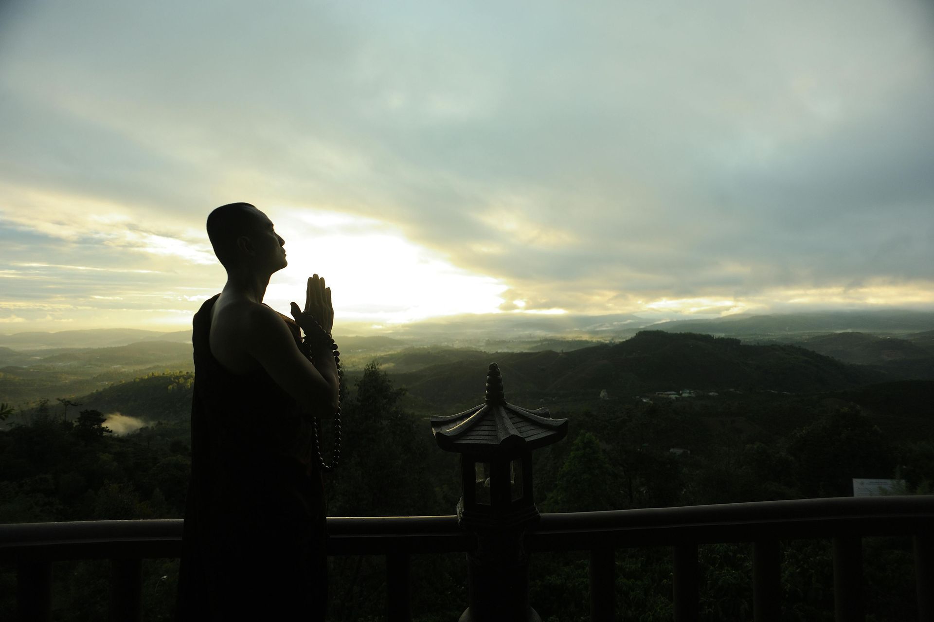 A man is standing on a balcony with his hands folded in front of a sunset.