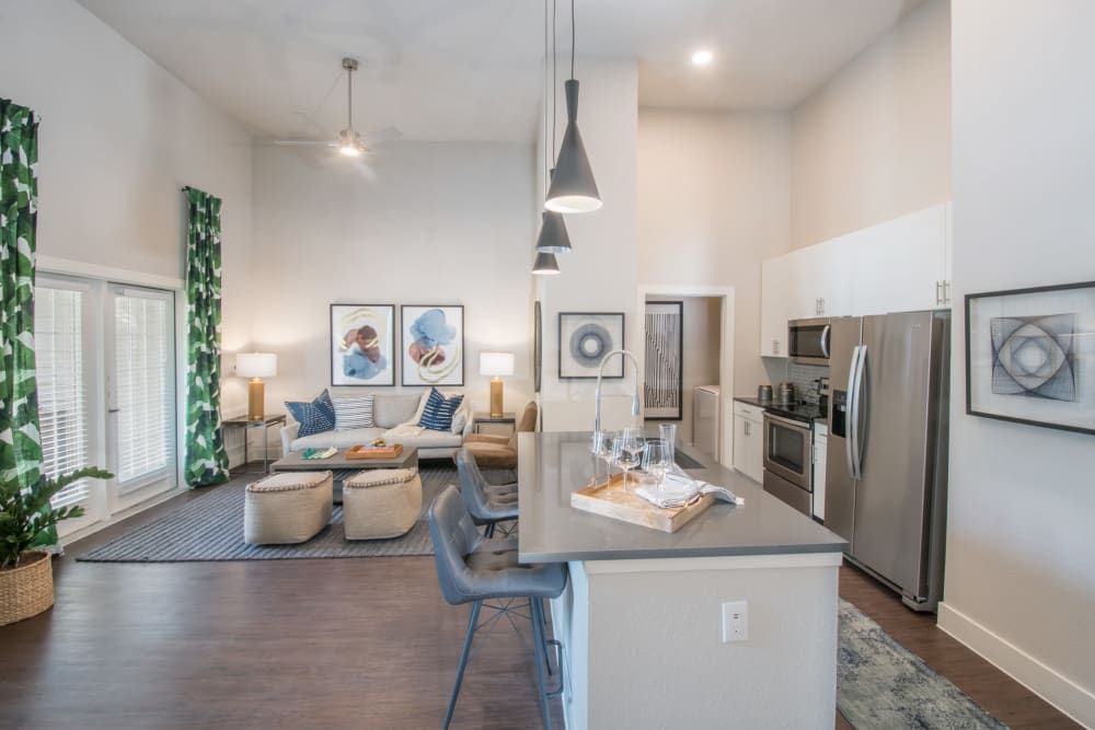 Modern apartment interior featuring an open-plan living room with a seating area that includes a gray sofa, armchairs, a patterned rug, and decorative green curtains. Two abstract paintings hang above the sofa. The living area transitions seamlessly to a kitchen with stainless steel appliances, white cabinets, and a gray countertop with bar stools. A chandelier and pendant lights add a contemporary touch to the space at Marquis Dominion in San Antonio, TX. 