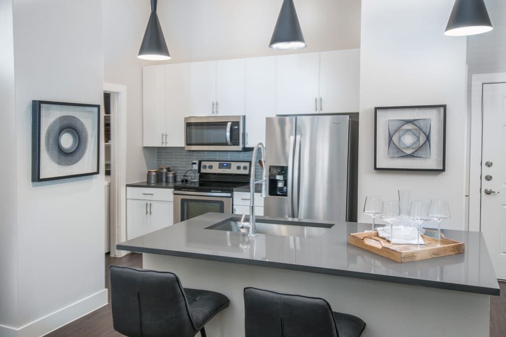 Modern kitchen interior with white cabinetry, stainless steel appliances, and dark gray backsplash. A kitchen island with a breakfast bar, two black leather stools, and pendant lighting above it is in the foreground. Decorative geometrical artwork hangs on the white walls, creating a chic and clean look at Marquis Dominion in San Antonio, TX. 