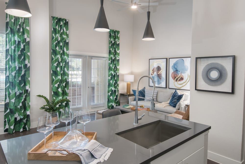 Modern kitchen interior with geometric green and white curtains, sleek gray countertops, and elegant hanging pendant lights. The room is styled with a wooden tray holding wine glasses on the island, abstract art pieces on the wall, and cozy pillows on a sofa in the background at Marquis Dominion in San Antonio, TX. 