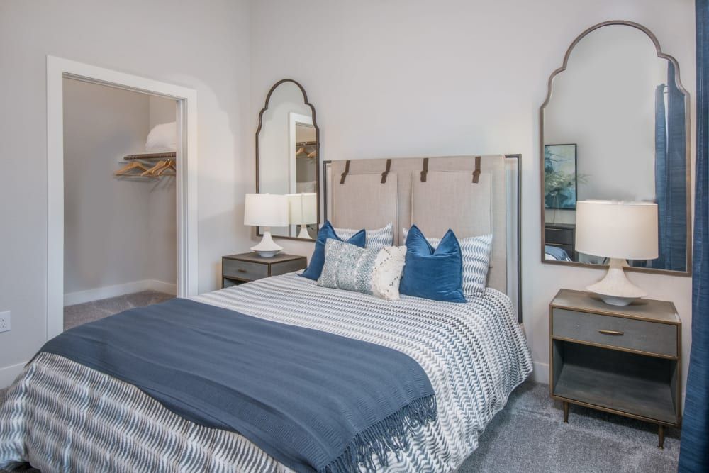 Modern bedroom interior with a neatly made bed featuring gray and blue striped bedding and several decorative pillows. Flanked by two nightstands with matching lamps, the bed has a tufted headboard against a pale gray wall. Above each nightstand is a decorative mirror with a similar design. To the left, an open closet with shelves is visible, and on the right, there is a wall-mounted artwork partially reflected in the mirror at Marquis Dominion in San Antonio, TX. 