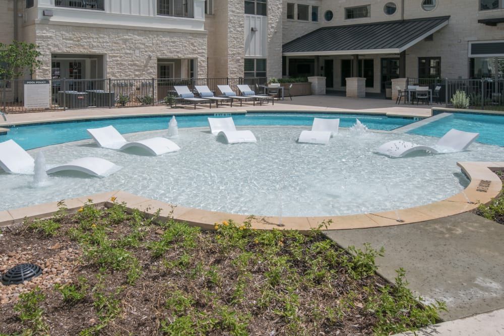 Outdoor swimming pool area with modern white lounge chairs partially submerged in the water, surrounded by a stone tiled patio and adjacent to a multi-storied residential building with several poolside seating arrangements at Marquis Dominion in San Antonio, TX. 