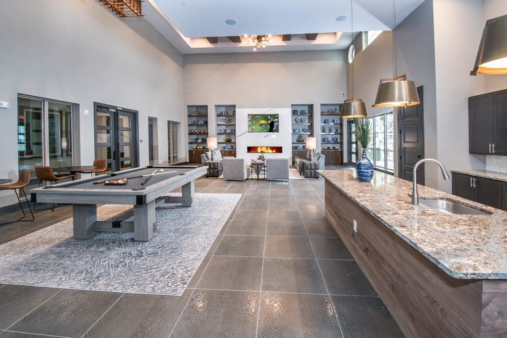 A spacious modern communal living area with high ceilings, featuring a pool table in the foreground, a kitchen island with a granite countertop, pendant lighting, and a comfortable seating area with a fireplace in the background. The room is styled with a neutral color palette and various decorative elements like shelves with books and ornaments, as well as potted plants at Marquis Dominion in San Antonio, TX. 