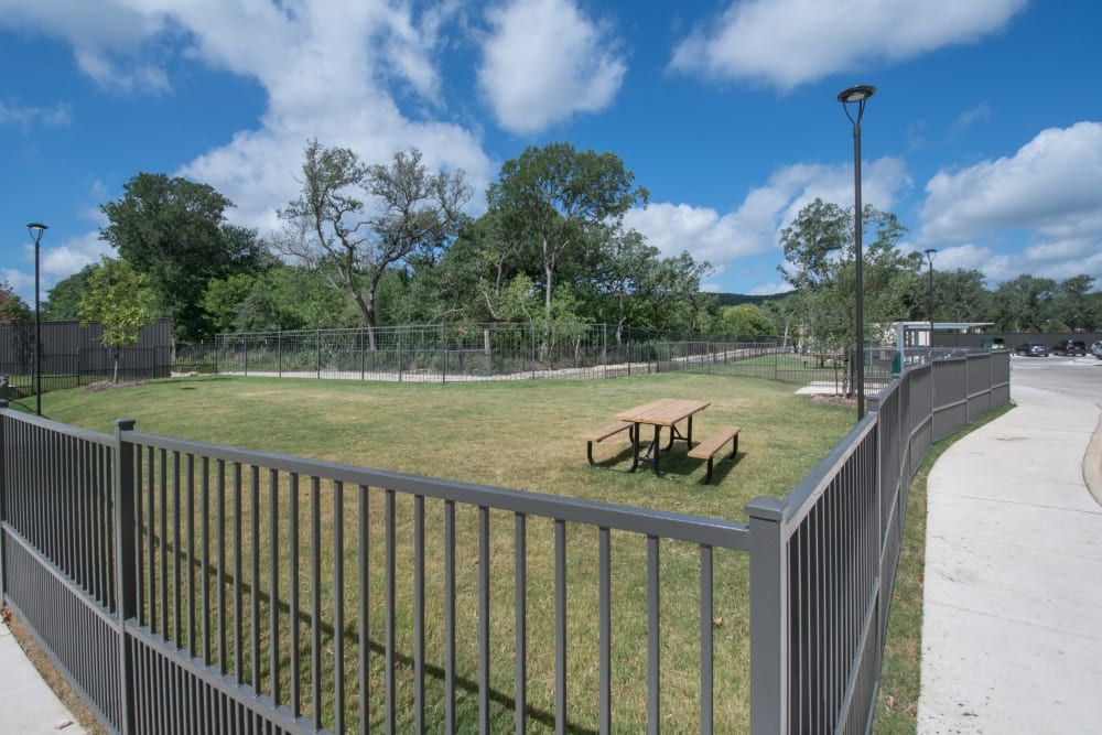 View of a well-maintained public dog park with a grassy area, surrounded by metal fencing, featuring a wooden picnic table and trees in the background under a blue sky with scattered clouds at Marquis Dominion in San Antonio, TX. 