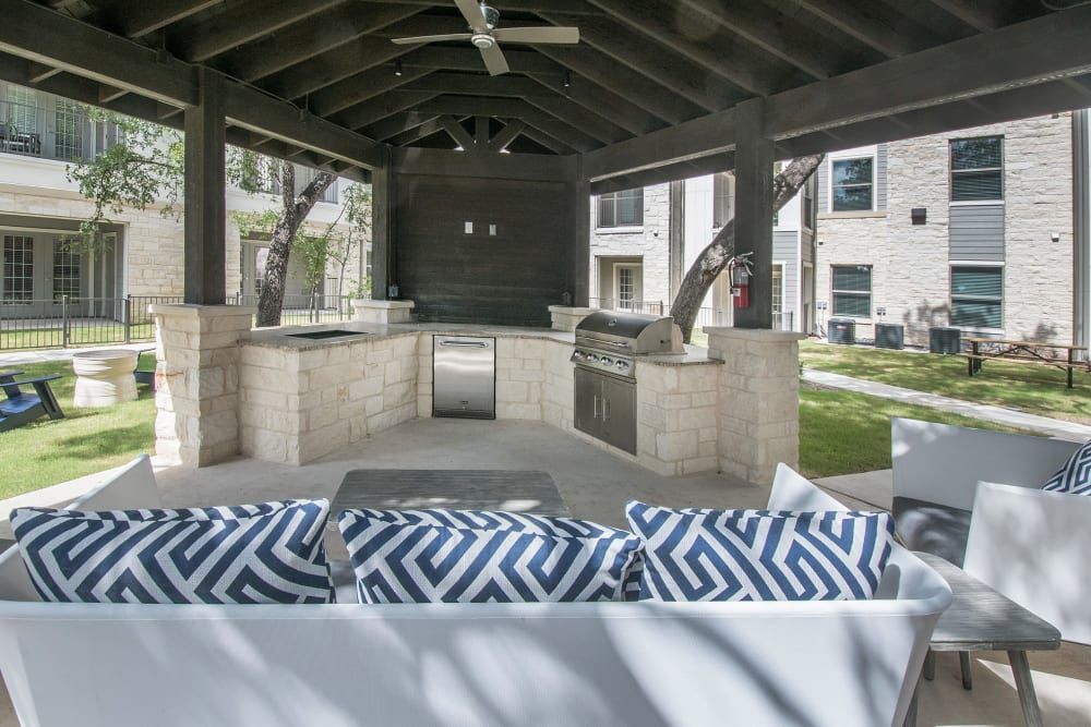 Open-air covered patio area with a modern outdoor kitchen, including a built-in grill and a countertop. The space features a white sofa with blue patterned cushions and a coordinated chair, set on a concrete floor. The background shows well-manicured grass and a multi-story building with stone veneer at Marquis Dominion in San Antonio, TX. 