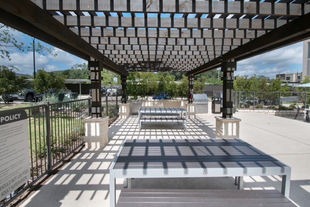 An outdoor pavilion featuring a grid-like wooden pergola casting patterned shadows over concrete flooring, with metal benches and tables, surrounded by a metal fence, lush greenery, and a clear blue sky above at Marquis Dominion in San Antonio, TX. 