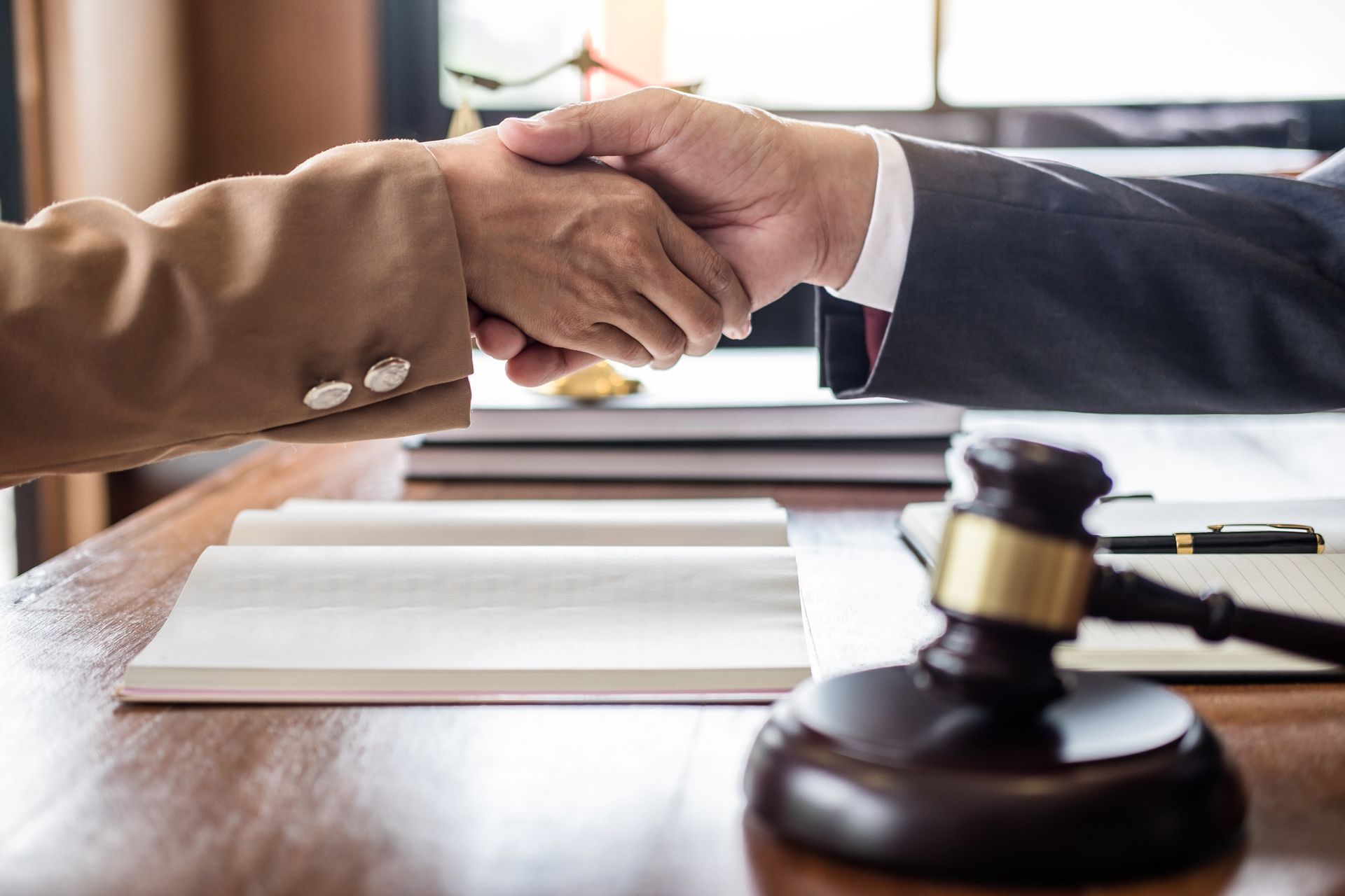 Two people shaking hands over a table with a gavel, legal books, and a pen.