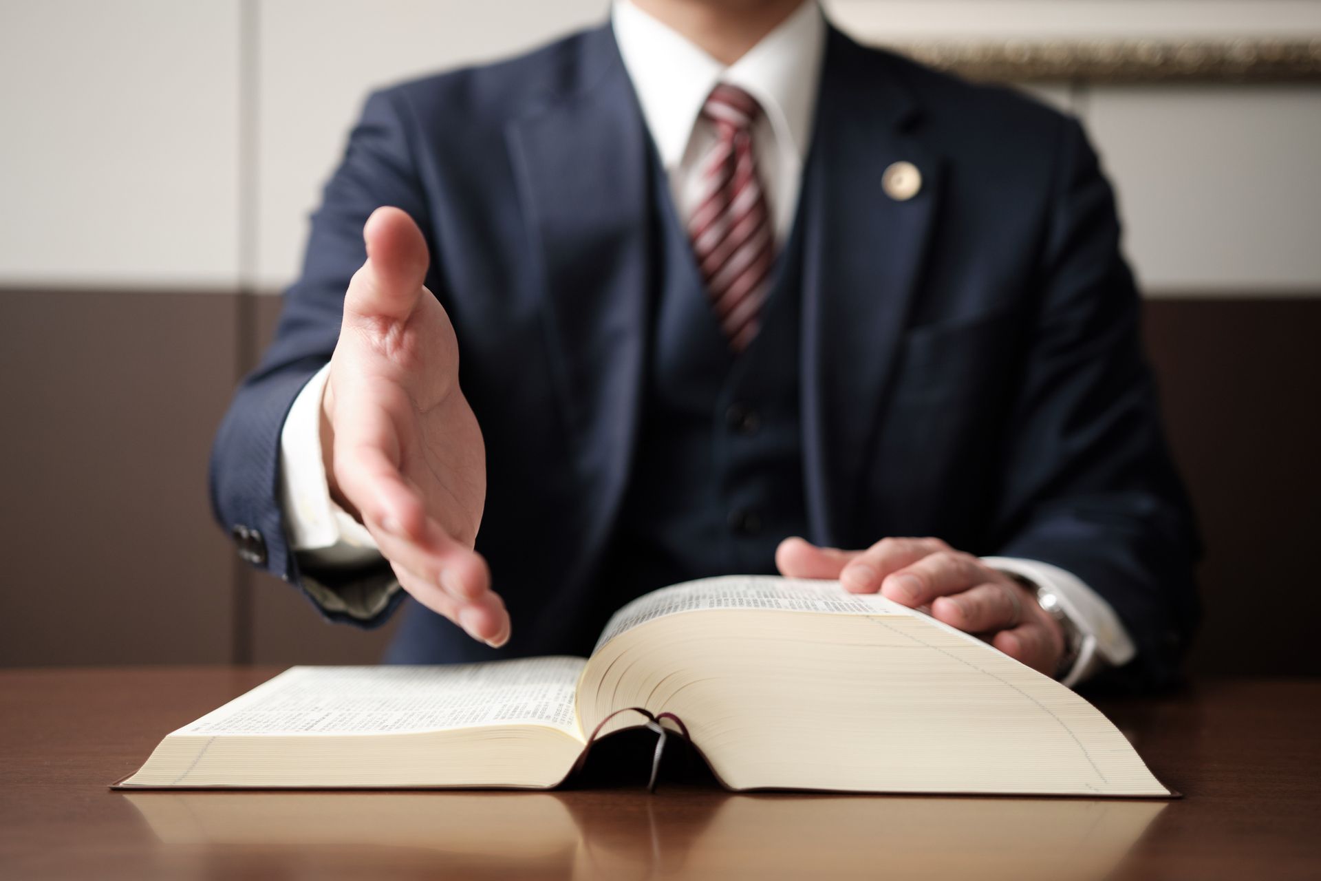 Man in suit extends hand for a handshake over an open book, likely in an office setting.