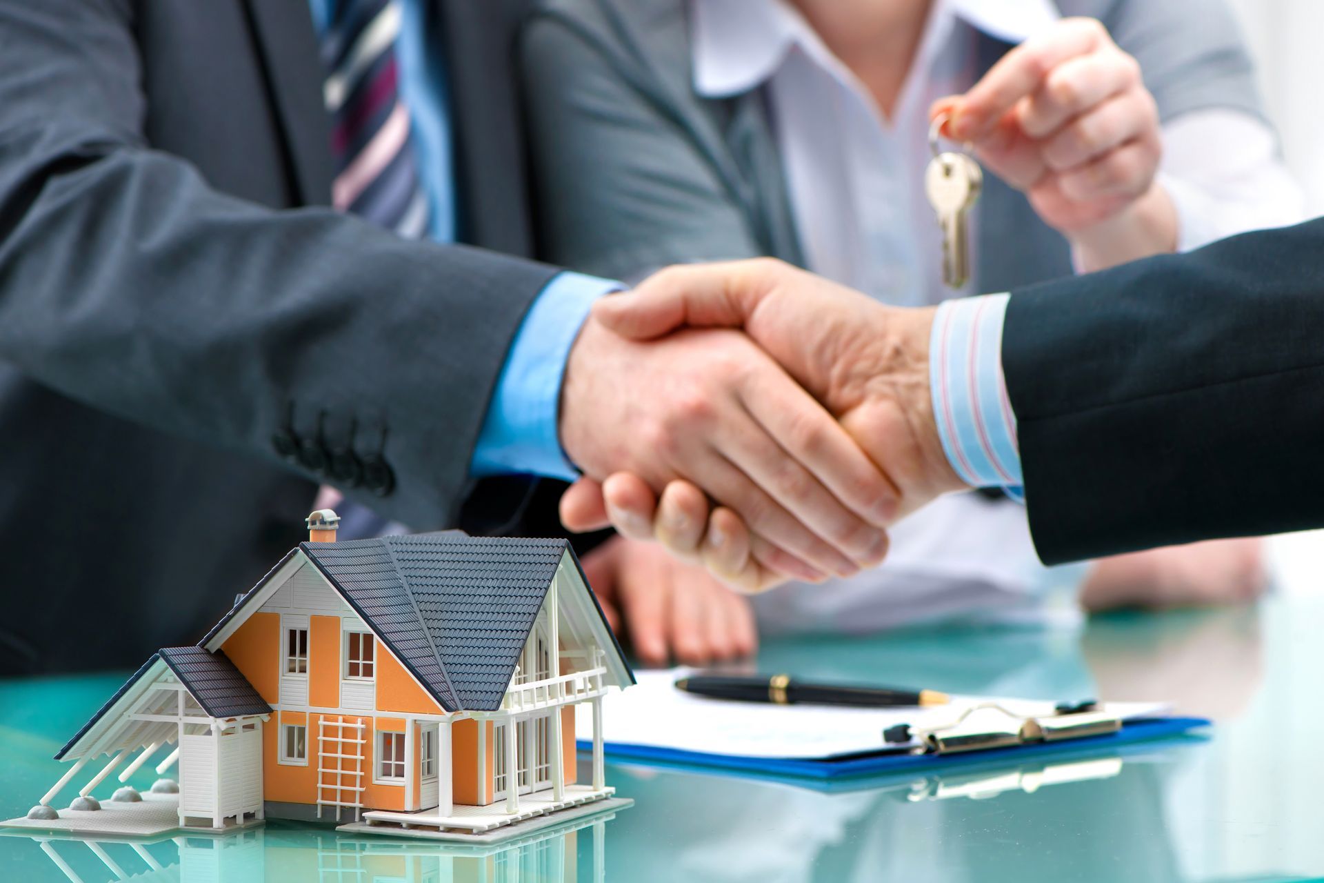 Handshake seals house deal. A person holds keys. Model house on a table next to a clipboard.