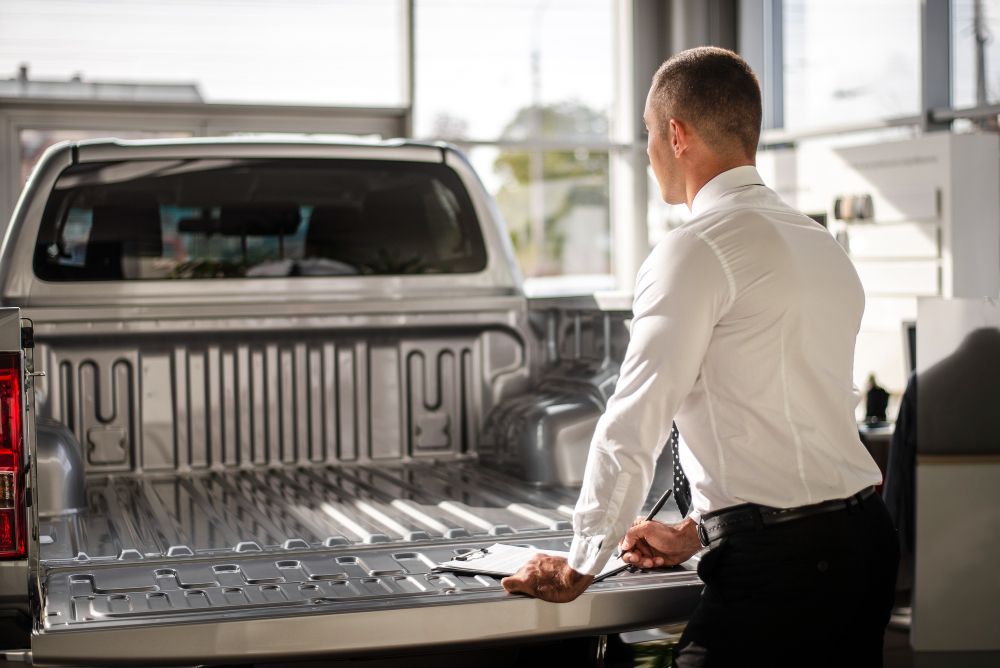 Man in white shirt looking at the bed of a silver pickup truck in a showroom.
