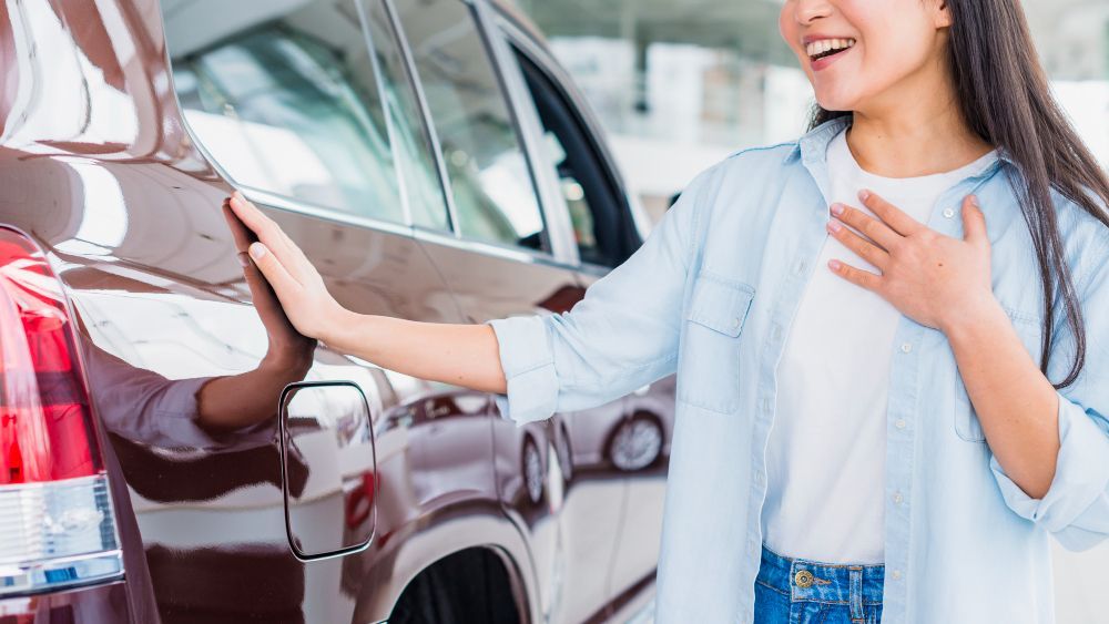 Woman admiring the side of a brown car, touching it with her hand and smiling. In a car dealership.
