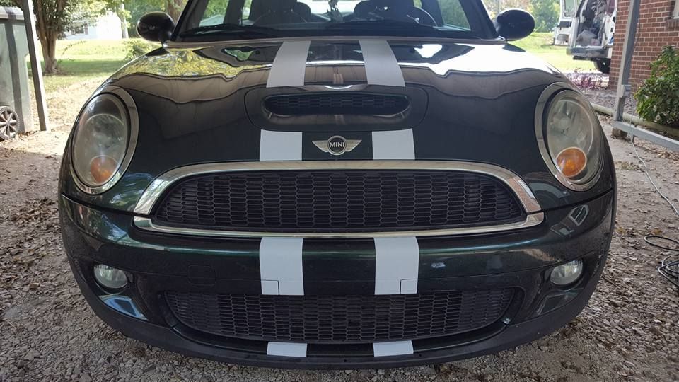 A black mini cooper with white stripes on the front is parked in a driveway.