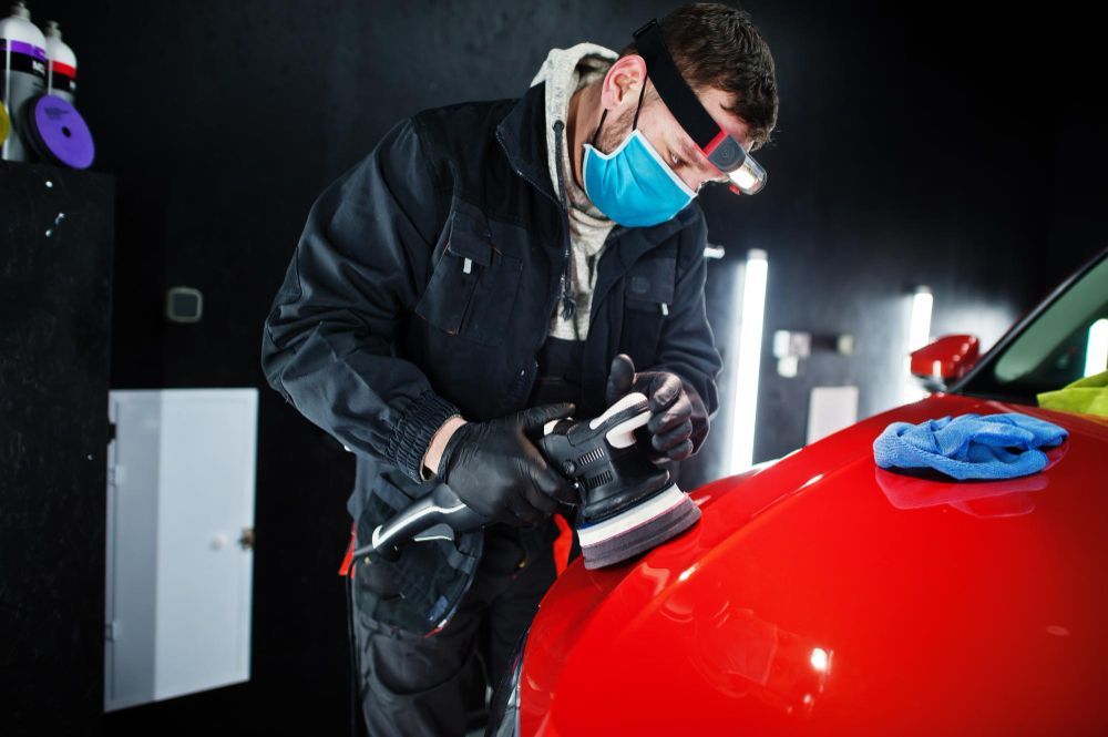 Person polishes a red car with a buffer, wearing a mask and safety glasses in a shop.