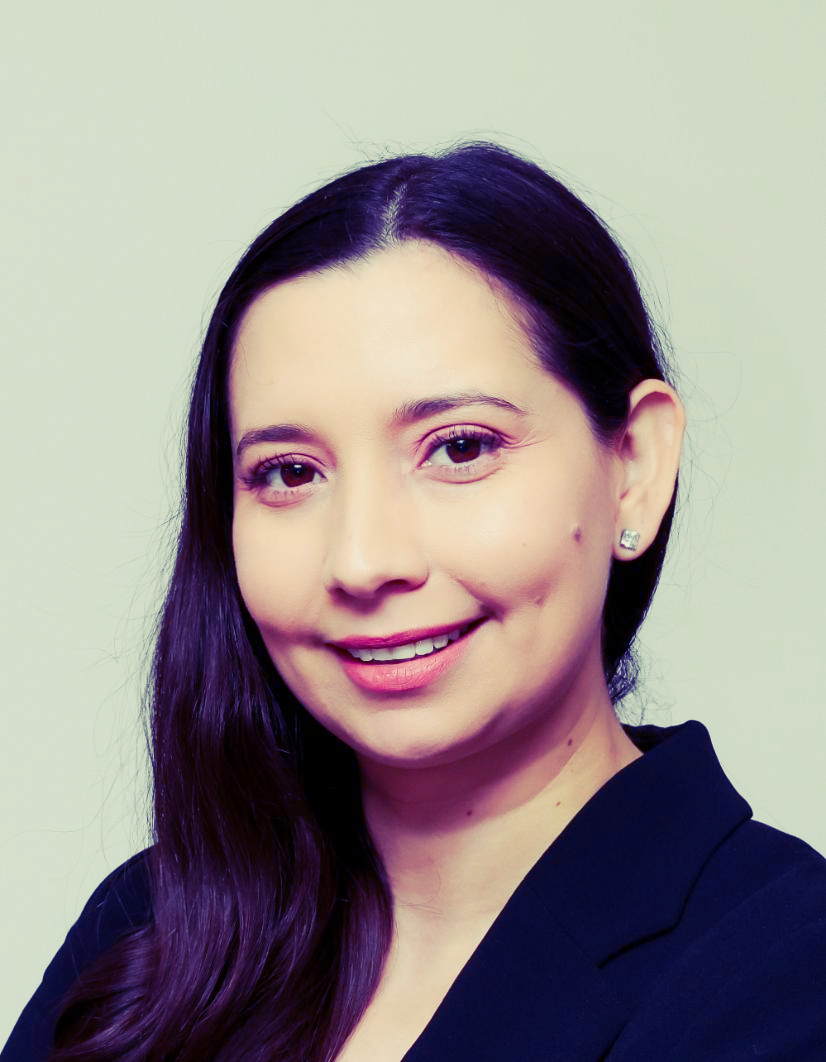 Woman with long dark hair, smiling, wearing a dark blazer, against a light background.