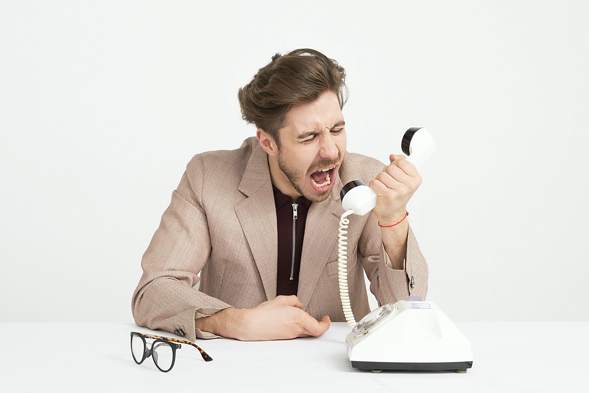 Man in tan blazer yelling into a white telephone. Glasses on table.