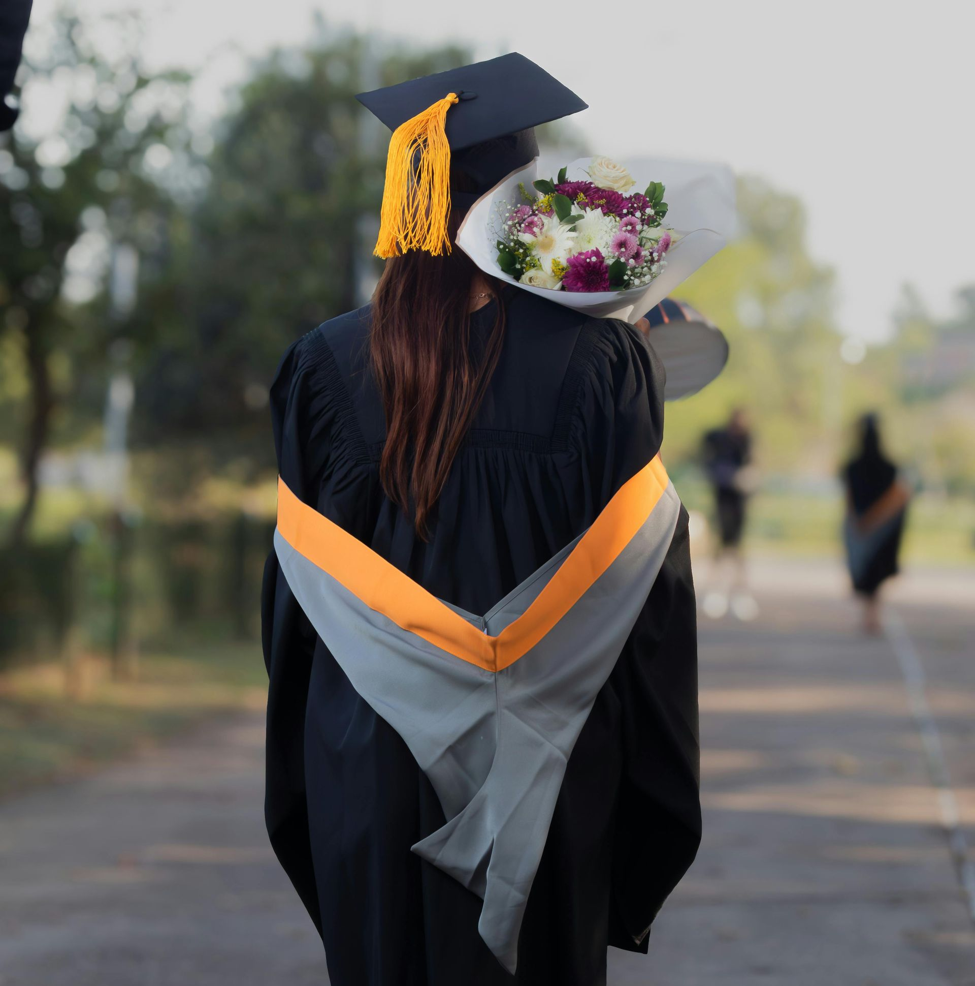 Graduation, person in cap and gown, holding flowers. Orange and gray sash, blurred outdoor background.