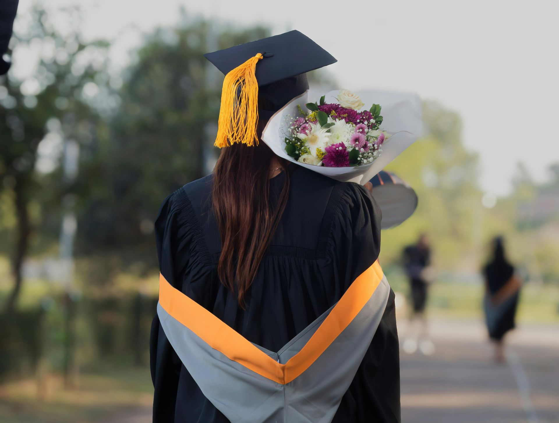 Graduation celebration: Person in cap and gown, holding flowers, walking on path.