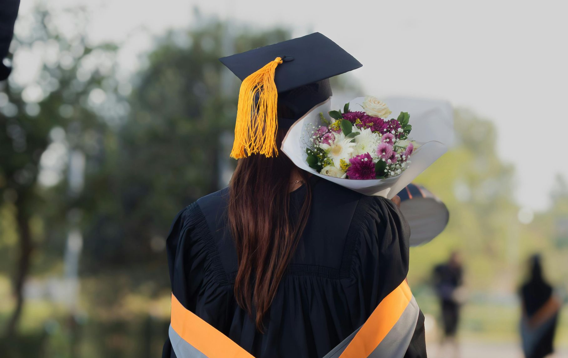 Graduation celebration: Person in cap and gown, holding flowers, walking on path.