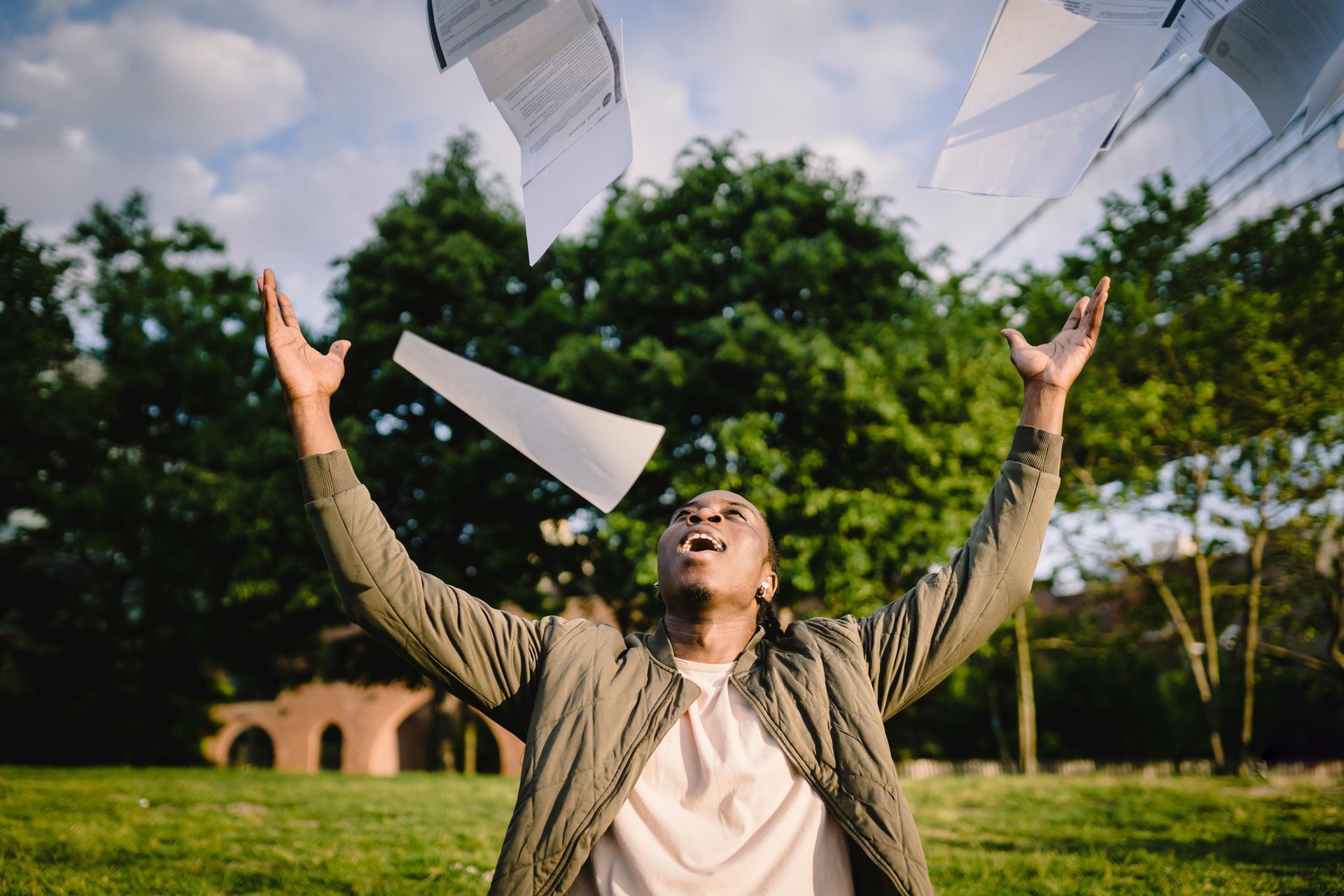 Person throwing papers up in the air with joyful expression in a green park.