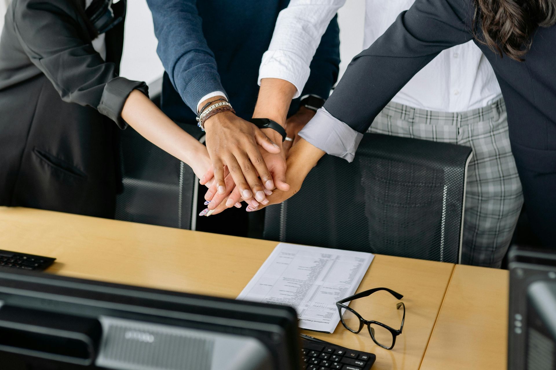 Hands of five people stacked together over a desk in an office setting.