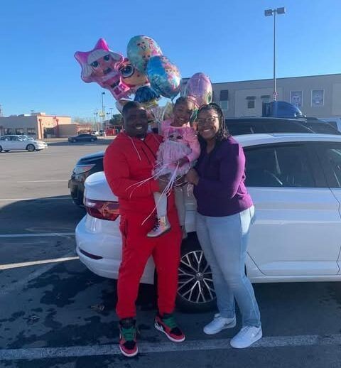 A man is holding a little girl and a bunch of balloons in front of a white car.