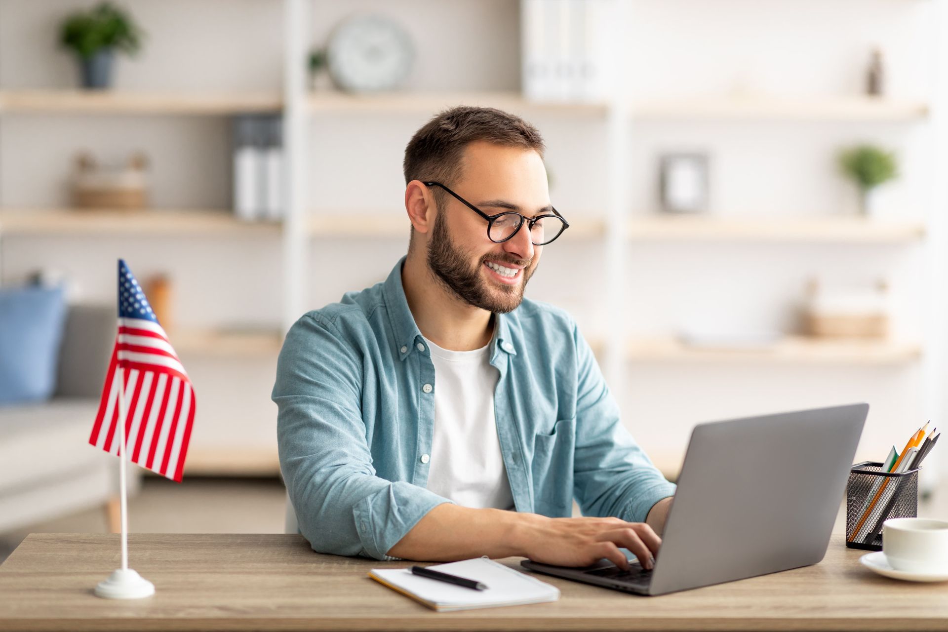 Man using a laptop at a desk with a small American flag in a bright home office