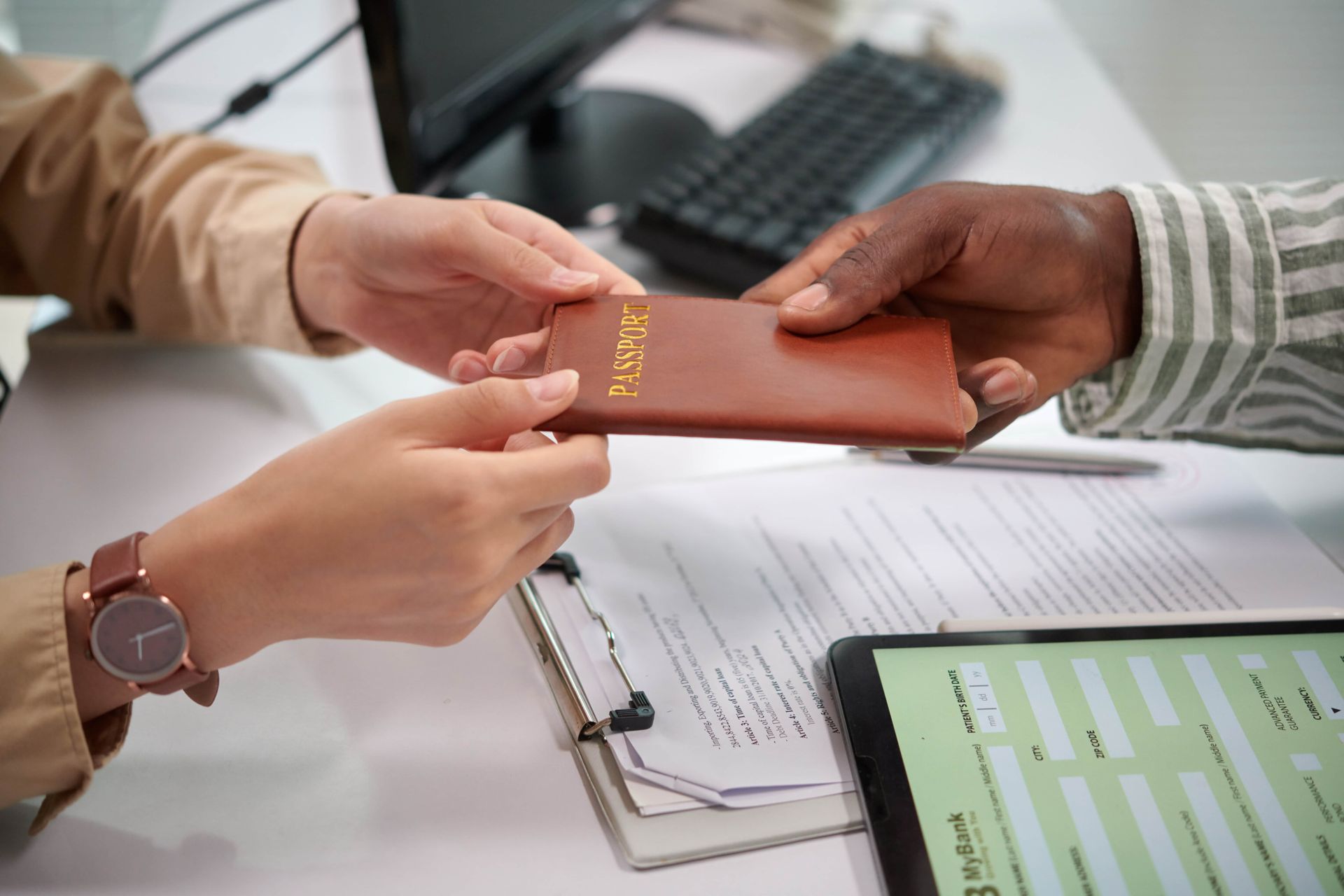 Hands exchange a brown passport over a desk with paperwork and a tablet visible