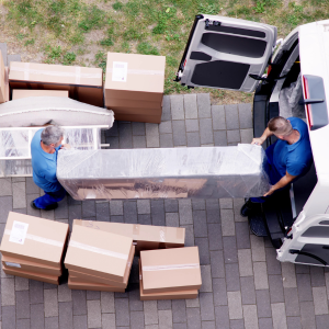 Two movers in blue shirts unload a long wrapped package from a white van onto a paved driveway.