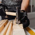 Gloved hands hammering a nail into a light wood plank on a workbench, with a measuring tape nearby