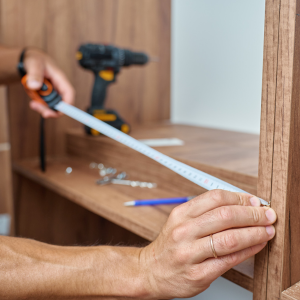 Measuring a wooden shelf with a tape measure while assembling furniture, with a drill nearby