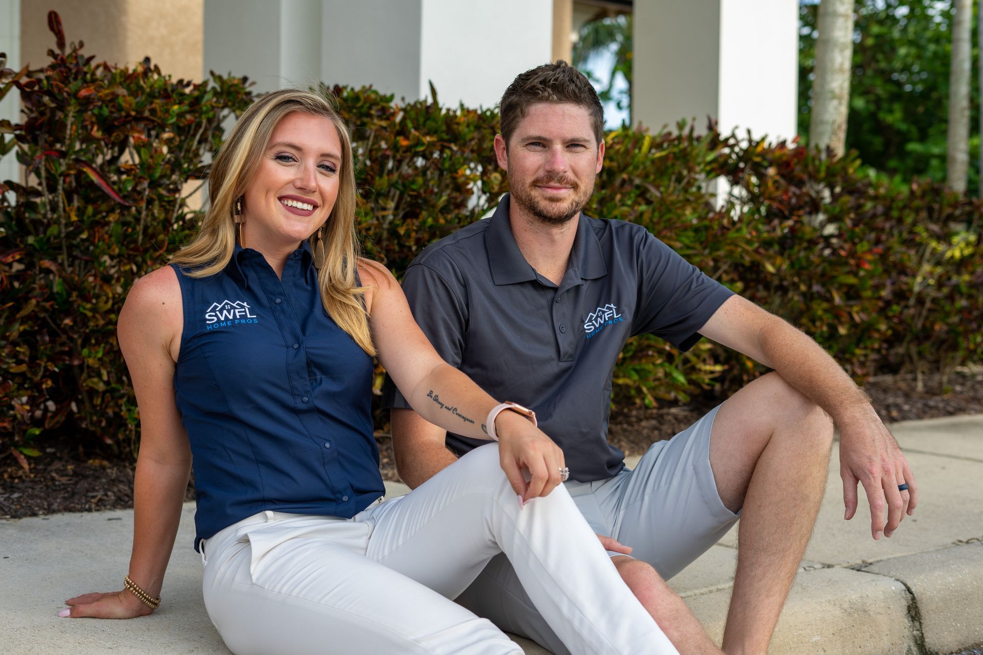 Woman and man sitting, smiling, wearing matching logo shirts, white pants; outdoor setting.