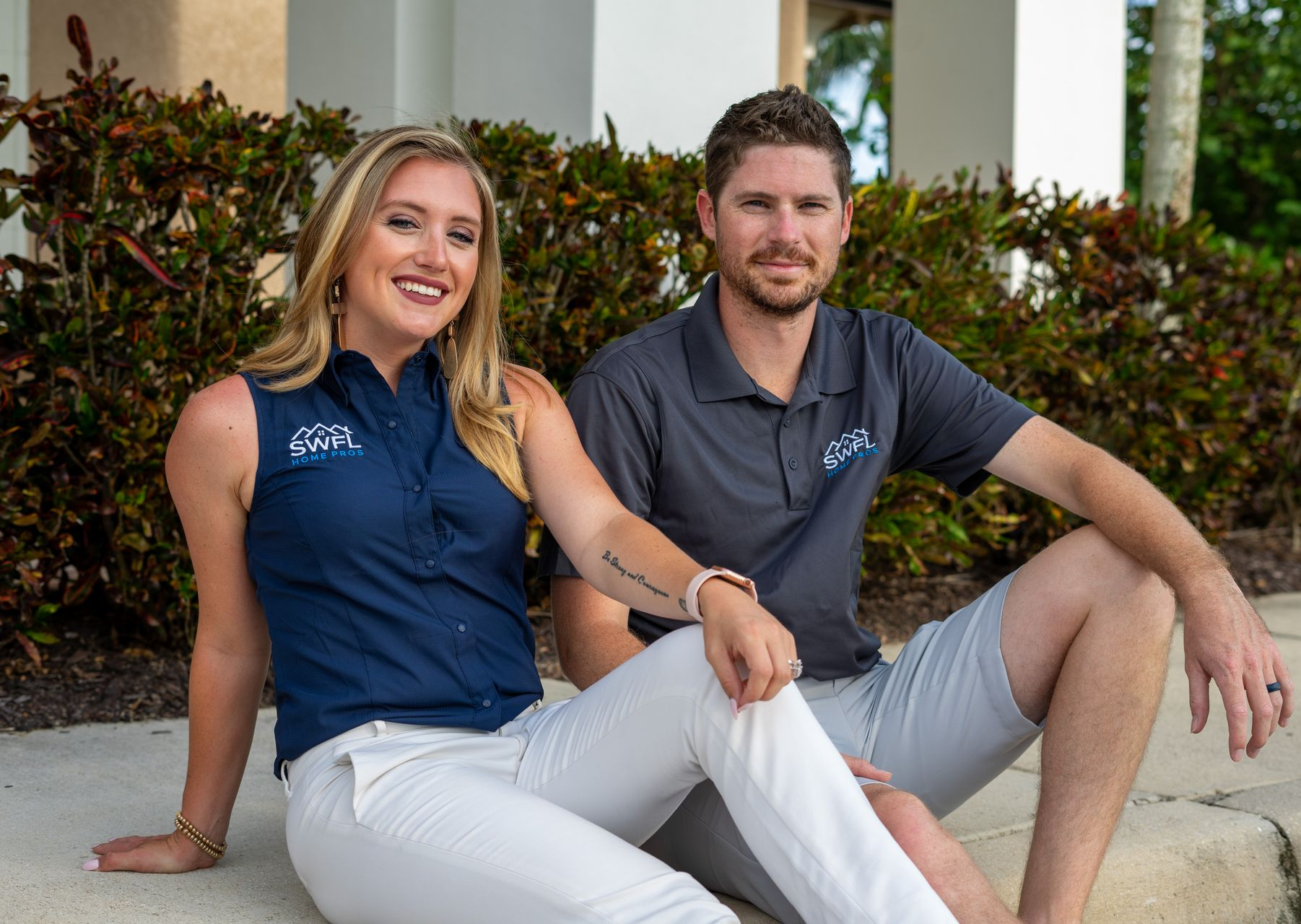 Woman and man sitting, smiling, wearing matching branded work shirts and pants outdoors.