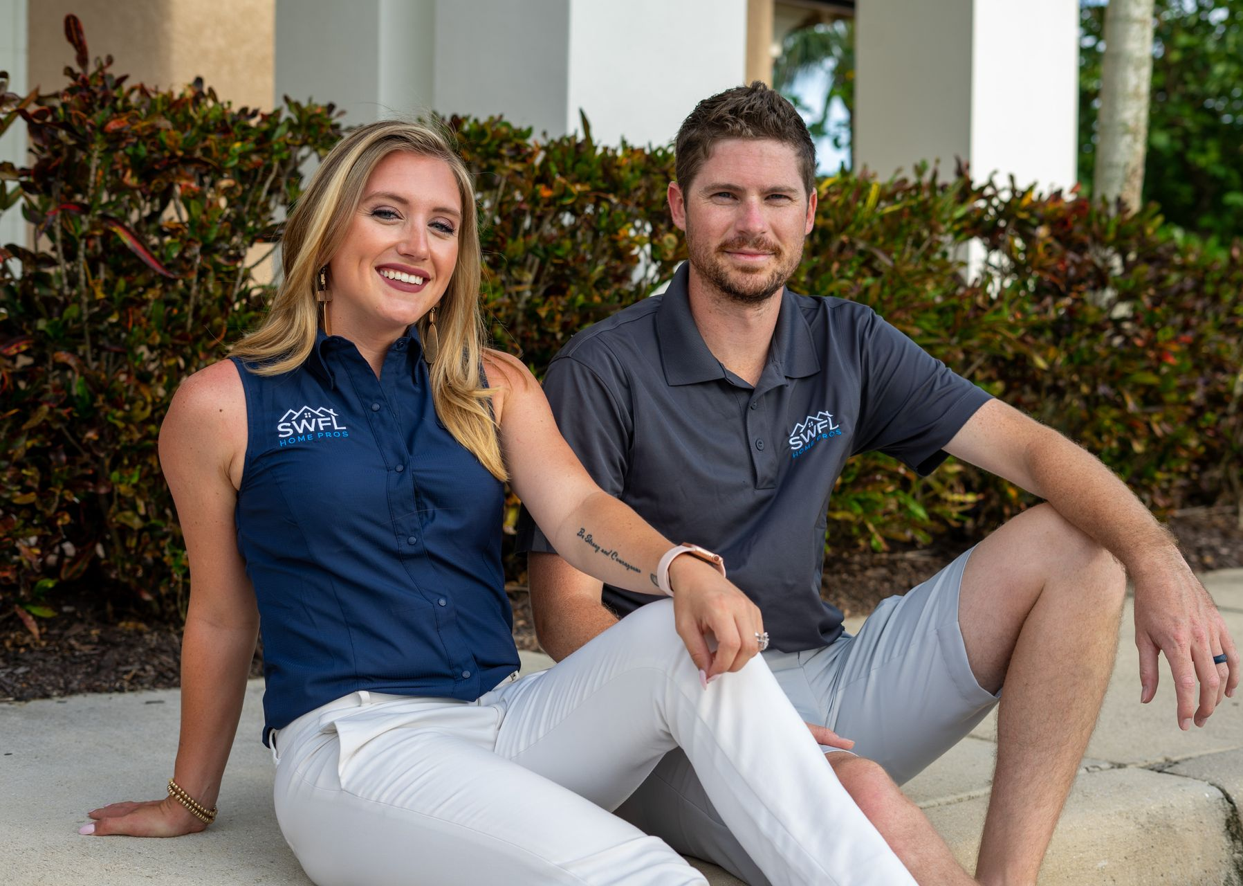Woman and man sit outside. Woman wears blue shirt and white pants, smiling. Man wears gray shirt and shorts.