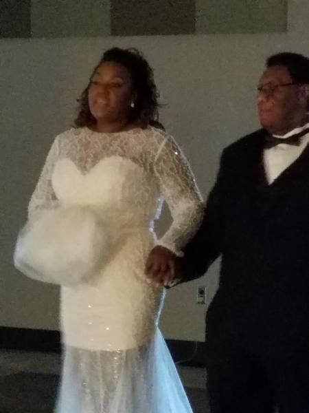 A couple in formal attire holding hands at an indoor event, with the woman in a white gown and the man in a black tuxedo.