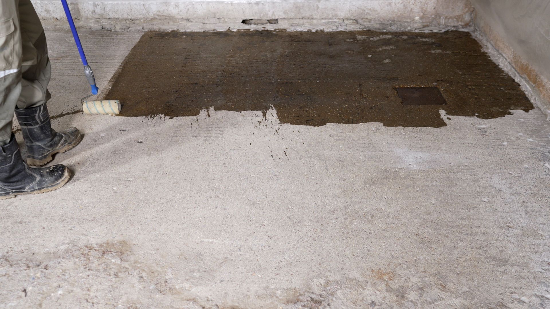Partially finished concrete floor with a dark patch being spread near a wall, and a worker’s boots at left