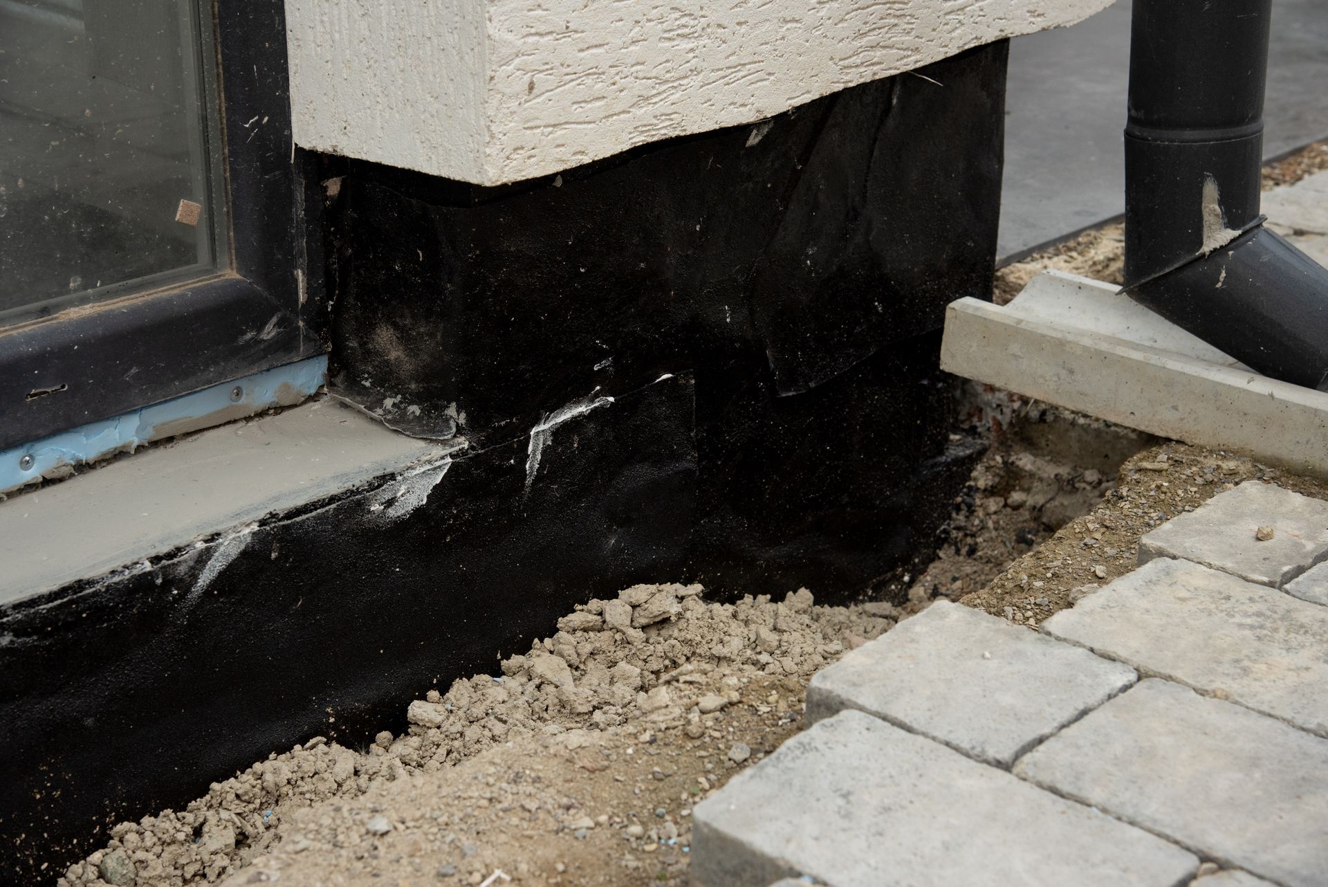 Damaged black foundation wall beside a building corner and paving stones, with exposed soil along the edge