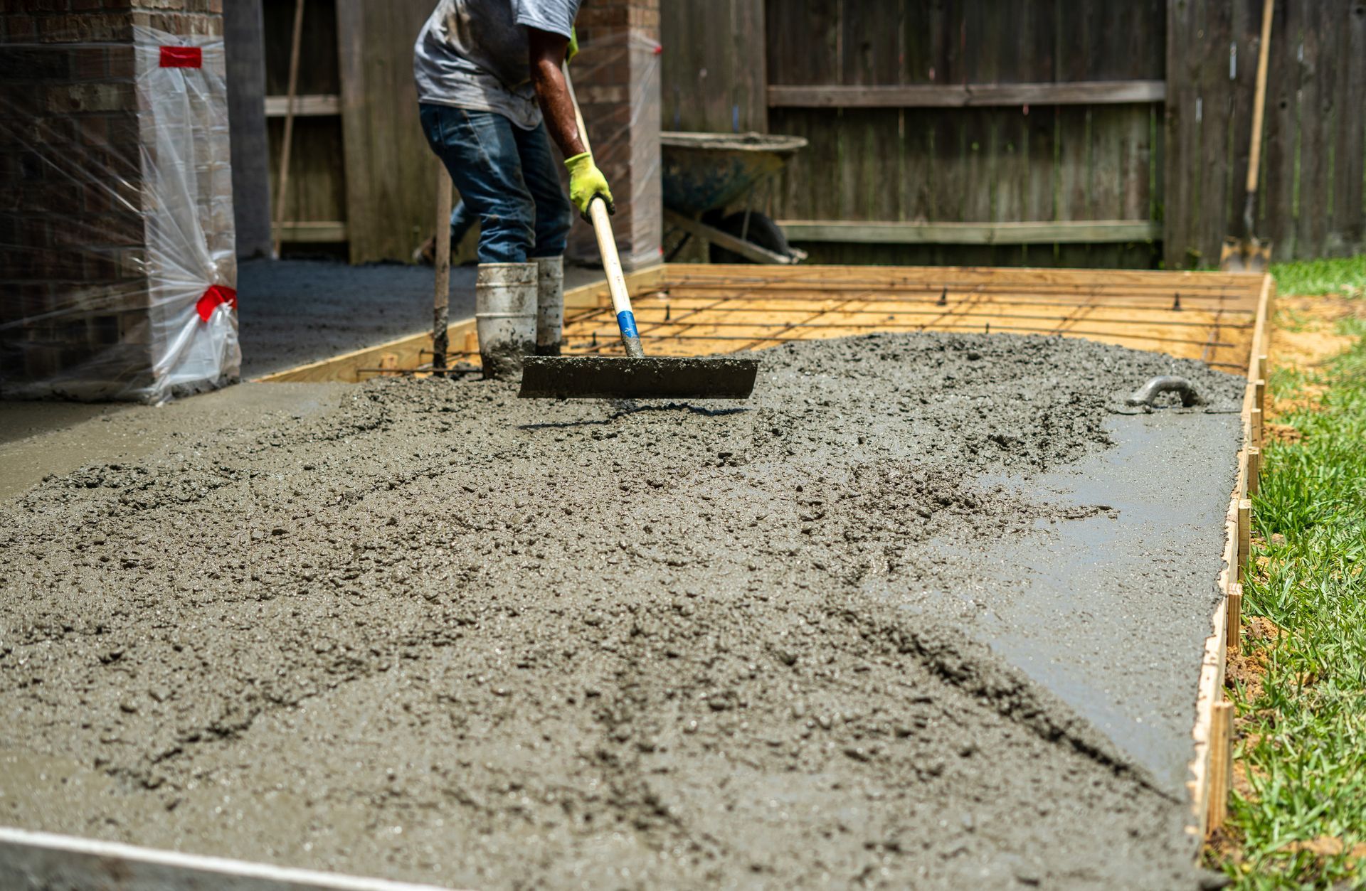 Worker smoothing wet concrete in a backyard patio area with a trowel and hose nearby