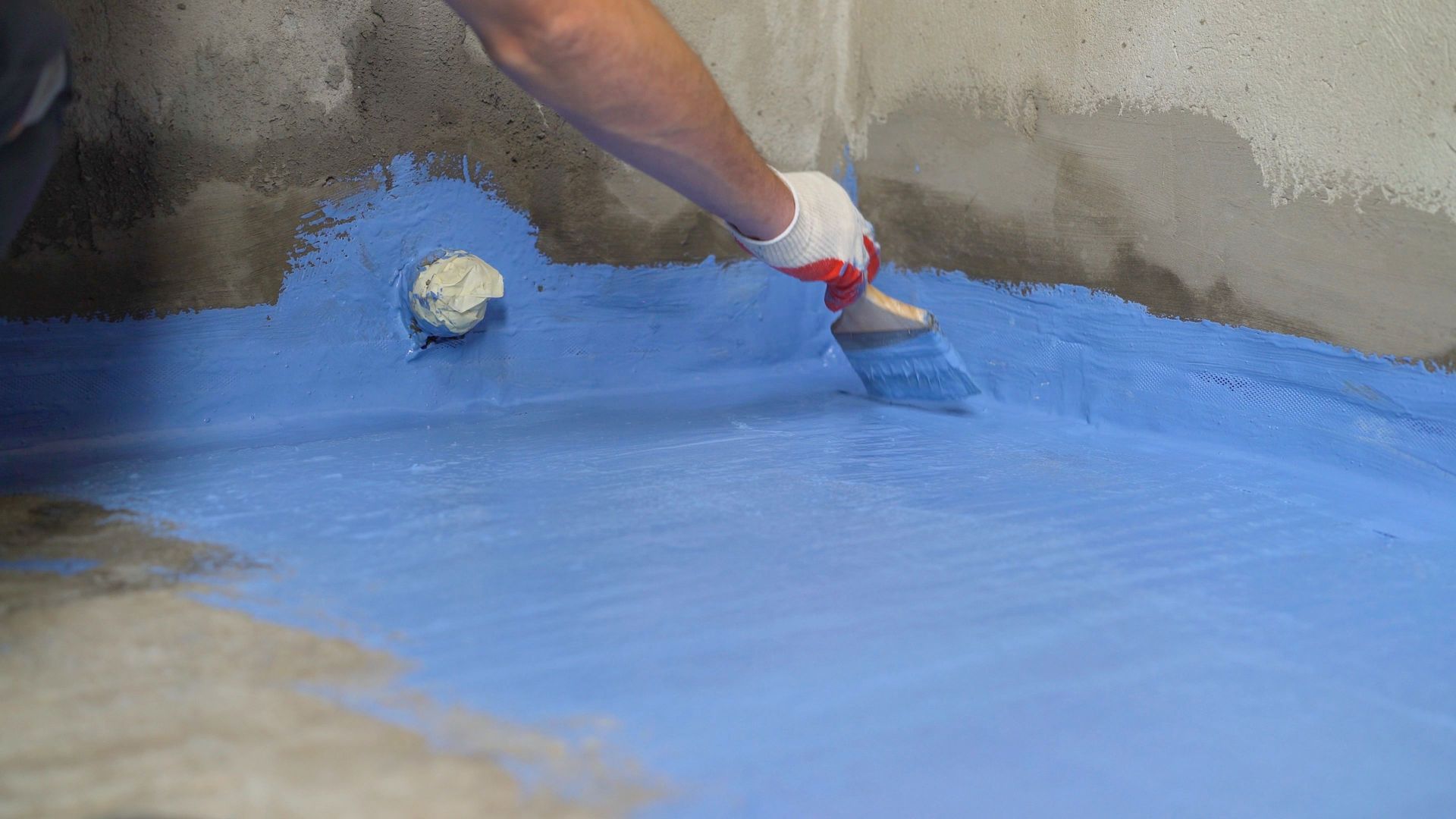 Worker applying blue waterproof coating with a brush on a concrete surface and wall corner